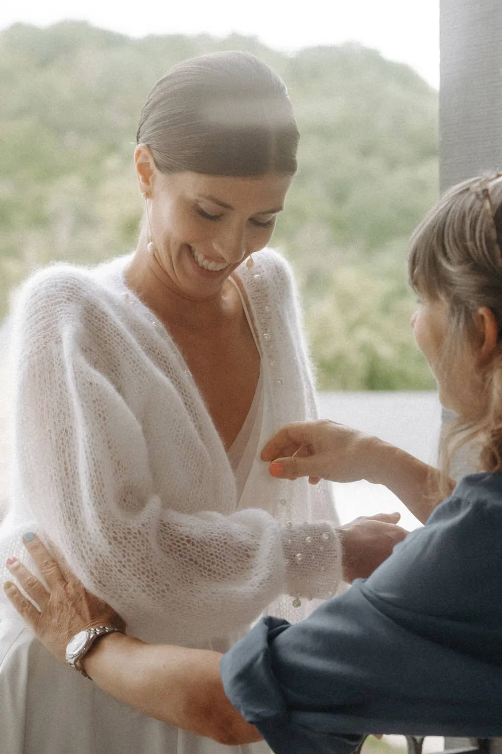 Braut wird beim Anziehen eines weißen Strickcardigans mit Perlenknöpfen unterstützt.