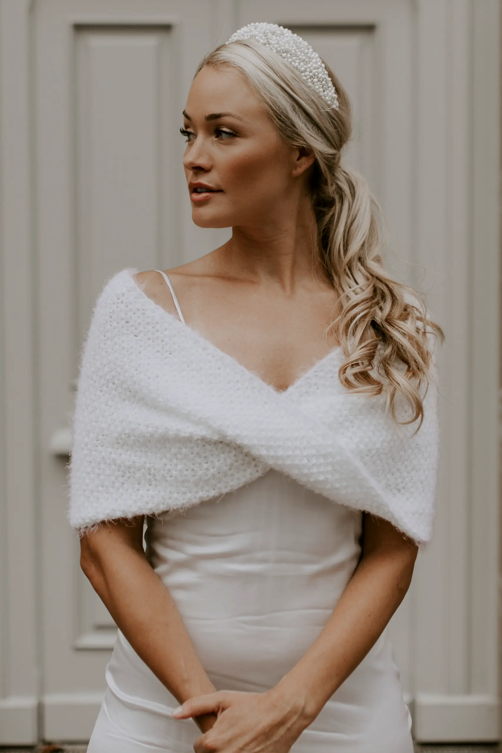 Eine Braut trägt den handgestrickten Brautloop PEARLY BERNIE in der Farbe Ivory über einem Brautkleid und ergänzt den Look mit einem Perlen-Haarreif.