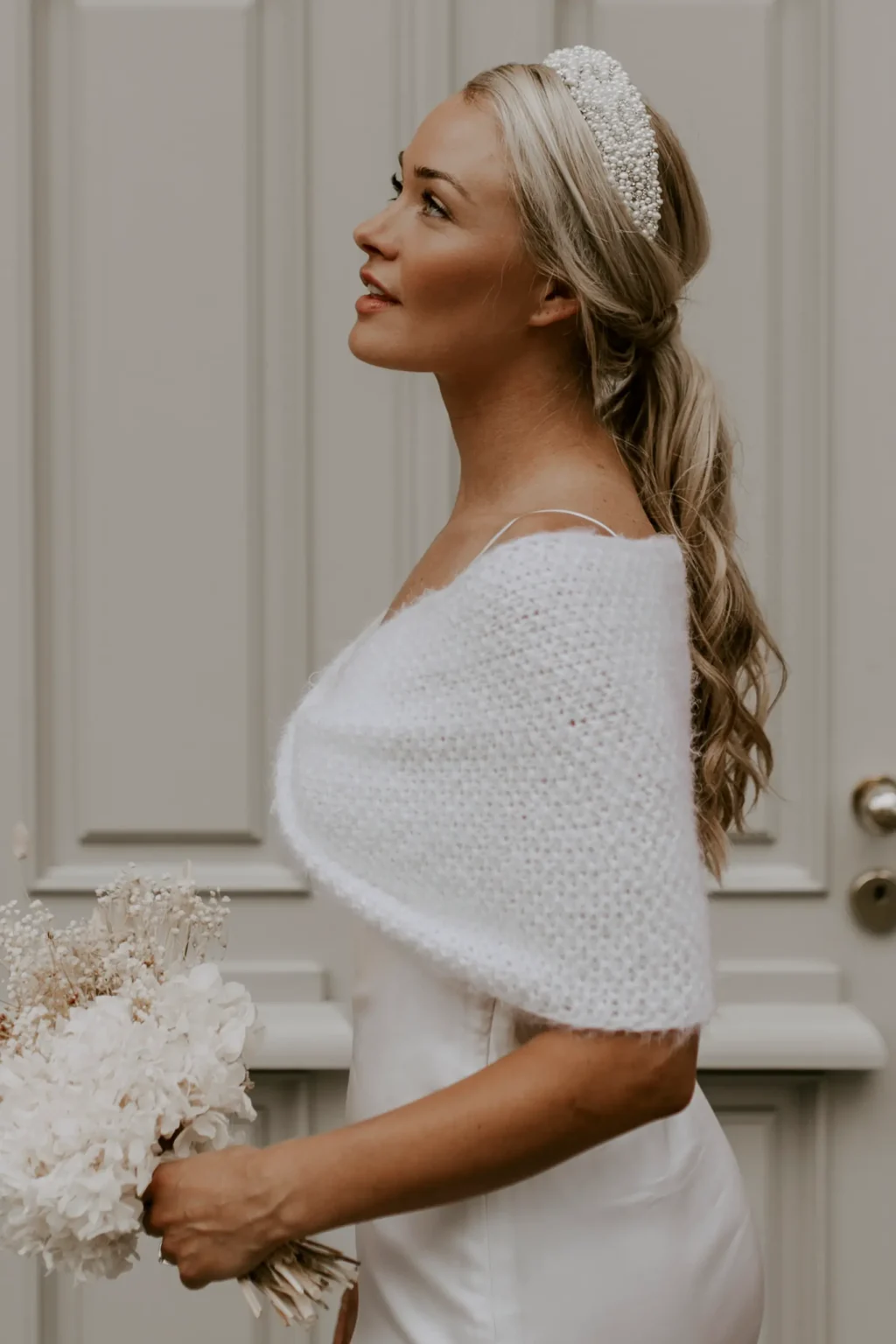 Eine Braut trägt den handgestrickten Brautloop PEARLY BERNIE in der Farbe Ivory über einem Brautkleid und ergänzt den Look mit einem Perlen-Haarreif.